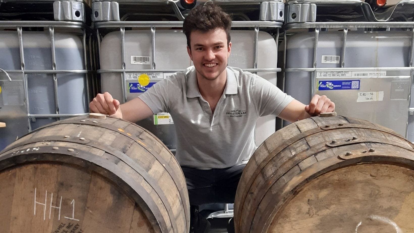 Shakespeare Distillery employee crouching and smiling behind two large wooden barrels